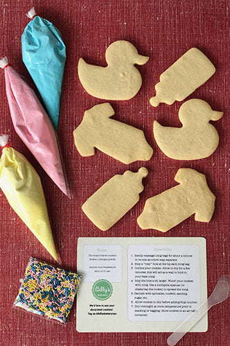 Smiley face emoji cookie decorating kit with plain and partially decorated round sugar cookies, yellow, white, and black icing bags, colorful rainbow sprinkles, and decorating instructions on a red textured background.