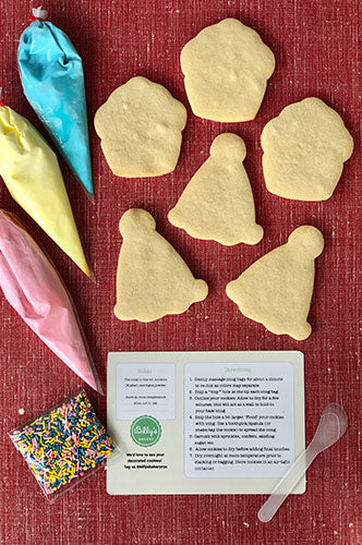 A red background with three un-iced heart-shaped cookies and three un-iced star-shaped cookies. Also pictured are three piping bags of yellow, pink, and blue icing, a bag of rainbow sprinkles, and a white instruction card.