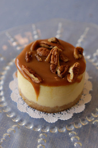 A slice of caramel pecan cheesecake with a layer of caramel on top and chopped pecans visible on the slice, served on a plate with a lace-like border.