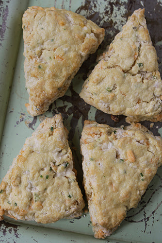 Four triangular cheddar chive scones on a light surface, with visible flaky layers and herbs sprinkled on top.