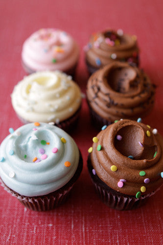A chocolate cupcake with chocolate frosting and decorative white sprinkles on top, placed on a cupcake holder with a floral patterned fabric underneath.