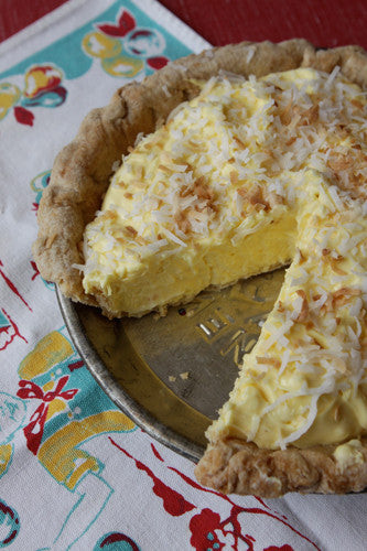 A coconut cream pie with a slice missing, topped with toasted coconut flakes, on a plate with a colorful patterned tablecloth in the background.
