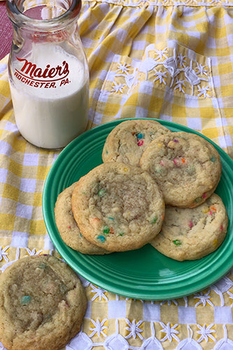 A green plate with chocolate chip cookies on it, placed on a yellow and white checkered tablecloth. There is a glass of milk in the background.