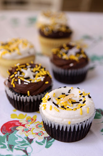 A collection of six assorted cupcakes, some with chocolate frosting and some with white frosting, all topped with yellow, black, and white sprinkles, and some with white star or stick decorations.