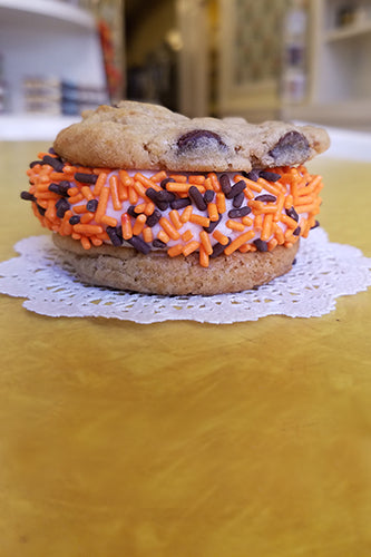 Chocolate chip cookie sandwich with orange and brown sprinkles on a light purple filling, placed on a white doily