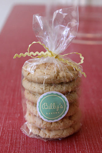 This inviting image features a tall stack of freshly baked, light golden-brown cookies, likely sugar or shortbread, neatly packaged in a transparent cellophane bag. The top of the bag is gathered and tied with a cheerful, wavy yellow ribbon, giving it a charming, homemade or artisanal feel. Adhered to the front of the bag is an oval-shaped sticker with a light green background and a darker green border. The sticker prominently displays the text "Billy's Bakery" in a stylish script font.