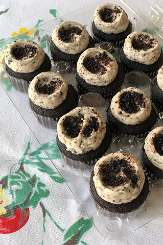 A dozen of chocolate cupcakes with vanilla buttercream frosting and cookie crumbles on top, displayed on a plate with a floral pattern.