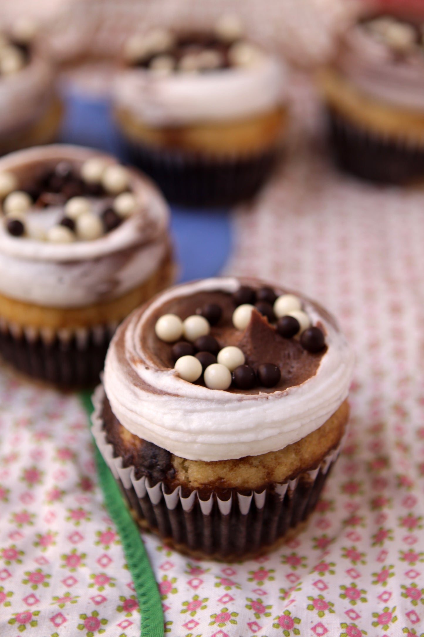 A photo of cupcakes with chocolate and vanilla batters, frosted with vanilla and chocolate buttercream and garnished with assorted chocolate pearls on a floral patterned surface.
