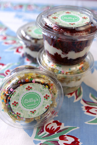 Clear plastic cups with various cupcakes inside, each cup has a different color and design lid, displayed on a table with a floral pattern.