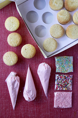 A set of ingredients for decorating cupcakes, including several cupcakes without frosting, a piping bag with white buttercream frosting, and a pouch of multicolored sprinkles, all laid out on a red surface.