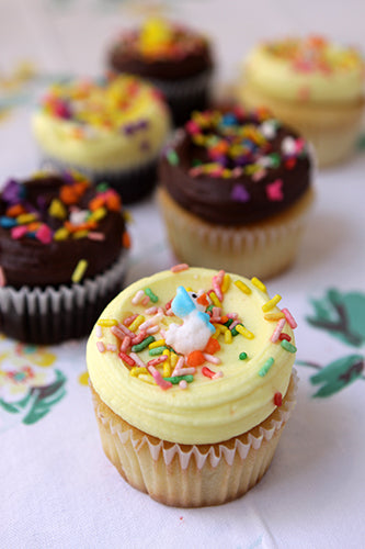 A selection of cupcakes with one frosted in yellow buttercream and topped with multicolored sprinkles, and another in dark chocolate frosting with a single white chocolate pearl on top. Both are on a paper liner with floral patterns.