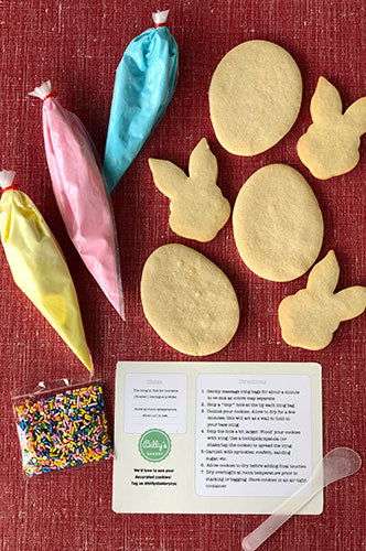 A red surface with three un-iced oval-shaped cookies and three un-iced bunny-shaped cookies. Also pictured are three piping bags of yellow, pink, and blue icing, a bag of rainbow sprinkles, and a white instruction card.