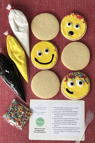 Emoji cookie decorating kit with round sugar cookies, yellow, white, and black icing bags, colorful sprinkles, and decorating instructions on a red background.