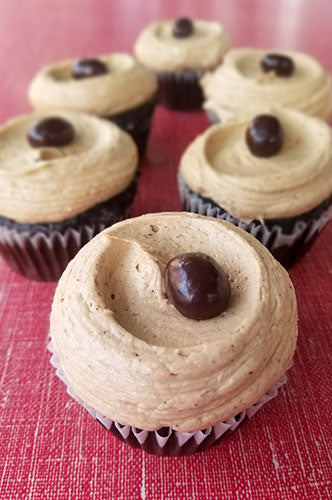 A photo of several espresso cupcakes with chocolate frosting and a chocolate-covered espresso bean on top, placed on a red surface.