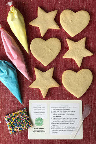 DIY cookie decorating kit featuring six plain sugar cookies in heart and star shapes, three piping bags of colorful icing (yellow, pink, blue), a packet of rainbow sprinkles, and an instruction card with a clear plastic spoon on a red background.