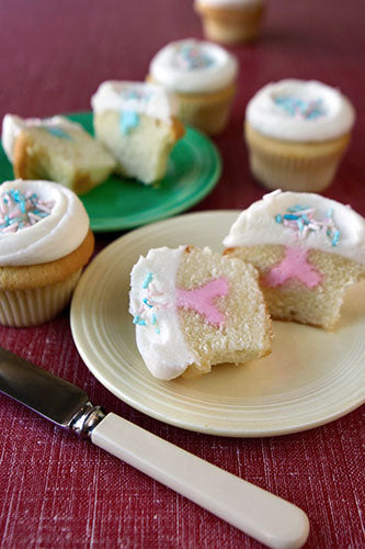 Close-up of a cut gender reveal cupcake revealing pink filling, with other frosted cupcakes and a knife in the foreground.