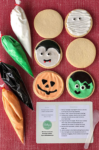 Overhead shot of Halloween-themed sugar cookies and decorating supplies. Four piping bags filled with white, green, black, and orange royal icing are on the left. On the right, decorated cookies include a mummy, a vampire, a jack-o'-lantern, and Frankenstein's monster, alongside two plain round cookies. A printed instruction sheet for decorating is at the bottom right.