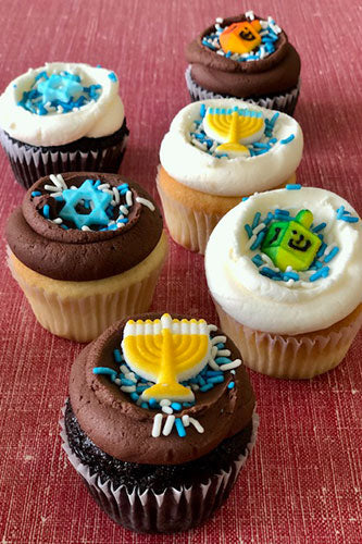 Six assorted Hanukkah-themed cupcakes, some chocolate and some vanilla, topped with white or chocolate frosting and edible menorahs, dreidels, or Stars of David, with blue and white sprinkles.