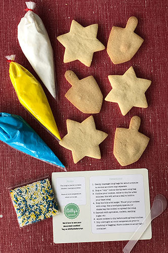 A variety of hand-cut sugar cookies in different shapes, with piping bags of royal icing and a mini spatula, displayed on a red surface with an instructional card.