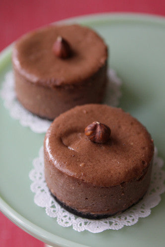Two small, round chocolate mousse desserts, each topped with a single hazelnut, resting on white lace doilies on a light green plate.