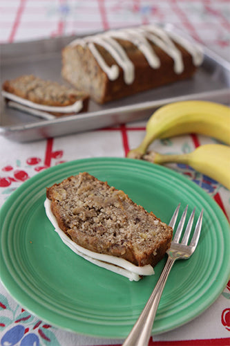 A loaf of banana cake with pineapple, spice, and pecans frosted on top, presented on a plate with a piece cut out to show the frosting. A whole banana and a piece of the cake are also visible in the background.