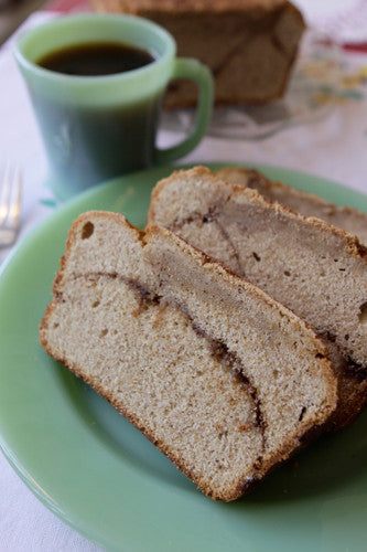 A loaf of cinnamon streusel coffee cake with a buttery, cinnamon crumb topping, cut into slices, served on a green plate with a cup of coffee and a piece of bread in the background.
