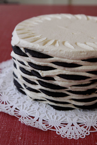 A round icebox cake with alternating black cookie and white cream layers forming a basket weave pattern on the sides, topped with smooth white cream and decorative piping, sitting on a white lace doily.