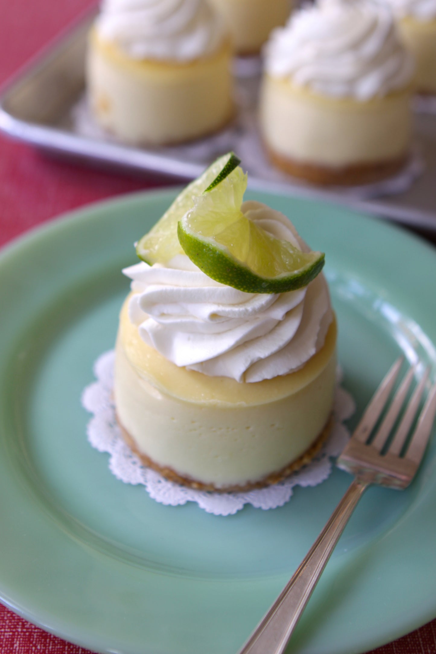 A piece of key lime cheesecake with a slice of lime on top, served on a green plate with a fork, with more cheesecakes in the background.
