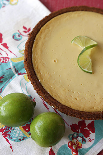 A key lime pie with a buttery graham cracker crust, garnished with a slice of lime, on a floral fabric with whole limes beside it.