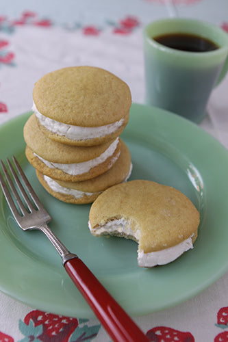 A plate with several maple cream whoopie cookies stacked on top of each other, with one whoopie partially eaten, and a red fork on the side. There is a cup of coffee in the background.