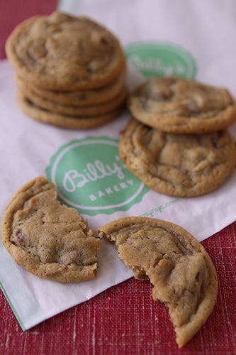 A pile of golden brown chocolate chip cookies on a white paper with 'Billy's BAKERY' logo, with two broken cookies in the foreground. The cookies appear soft and slightly chewy, with visible chocolate chips.