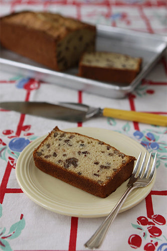 A loaf of maple pecan bread in a pan, cut in half, with a piece on a white plate with a fork, all on a red and white checkered tablecloth.