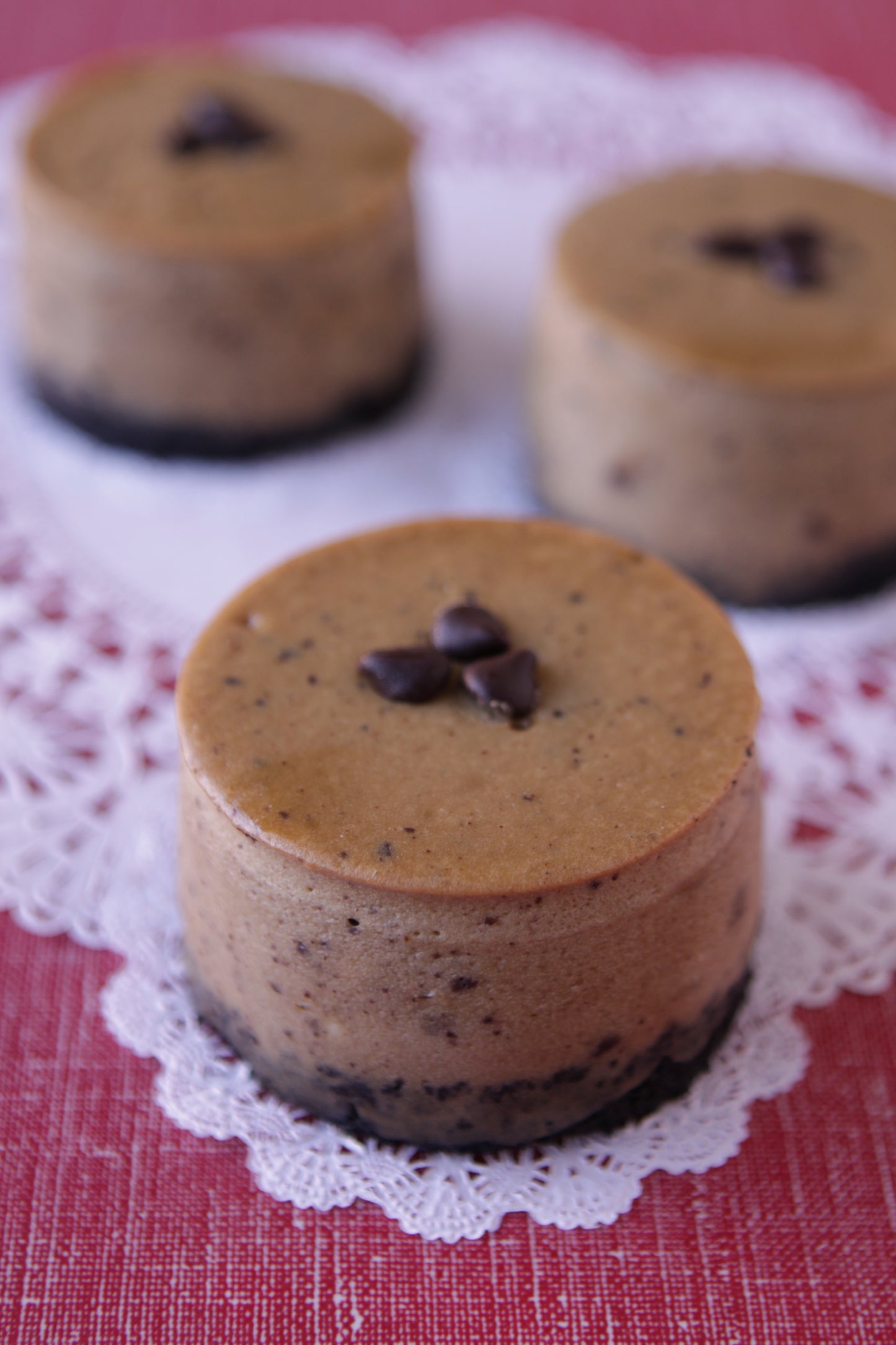 Three individual chocolate cheesecakes with chocolate chip toppings on a white doily over a red textured surface.