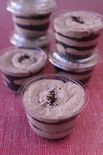 Close-up of multiple individual servings of Chocolate Cheesecake Trifles in clear cups, showcasing distinct layers of creamy light-colored cheesecake filling, dark chocolate, and a swirl of whipped cream topping garnished with chocolate crumbs