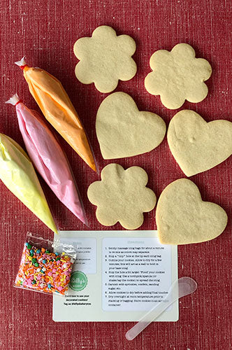 A Mothers Day themed cookie decorating kit featuring six plain sugar cookies in heart and flower shapes, three piping bags of colored icing (yellow, pink, and orange), a bag of colorful sprinkles, and an instruction card with a clear plastic spoon on a red textured background