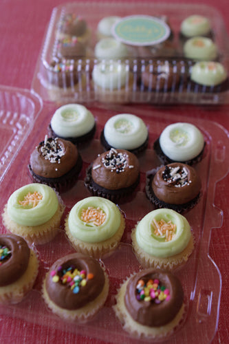A clear plastic container filled with assorted mini cupcakes, including chocolate frosted with sprinkles, and vanilla cupcakes with light green and white frosting, some topped with sprinkles.