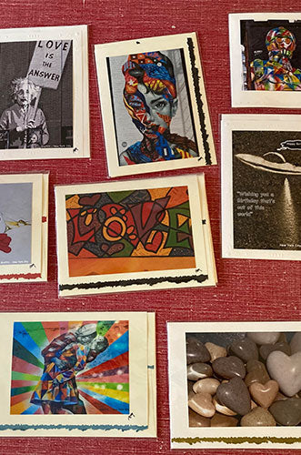 A collection of framed artwork and photographs arranged on a reddish-brown surface, including a black and white photo of a person holding a 'LOVE IS THE ANSWER' sign, colorful abstract art, a stylized portrait, and a photo of smooth stones and heart-shaped rocks.