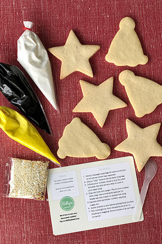 Six un-iced sugar cookies in shapes of stars and pointed party hats, arranged on a red textured surface with three piping bags (white, black, yellow icing) and a packet of white sprinkles, accompanied by decorating instructions.