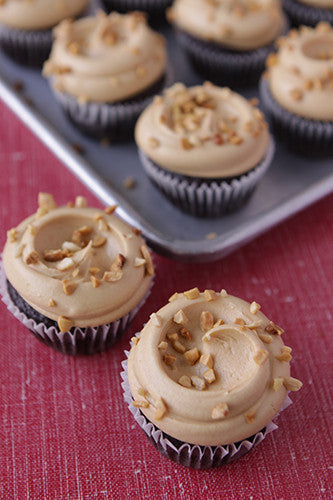 Chocolate cupcakes with creamy peanut butter frosting, topped with chopped peanuts, on a baking tray and a red textured surface.