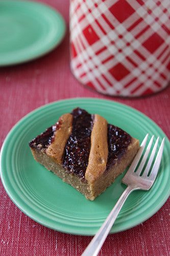 A peanut butter and jelly bar on a green plate with a fork, a red and white checkered cup in the background.