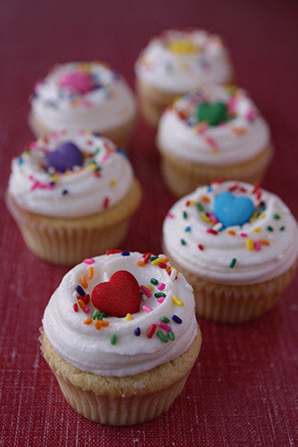 A group of cupcakes with white frosting and multicolored sprinkles, garnished with a heart-shaped decoration in various colors.