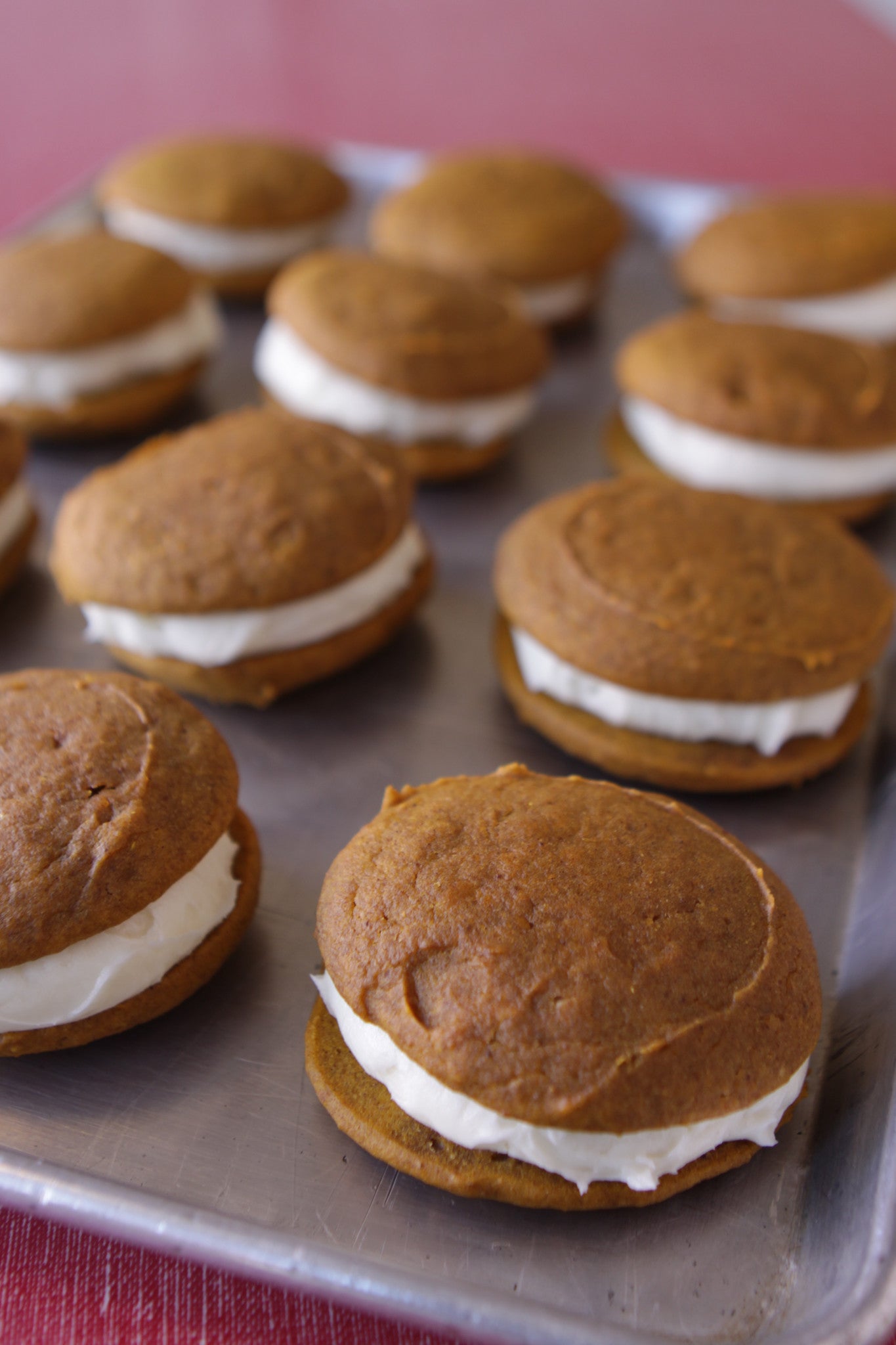 A tray of pumpkin-colored whoopie pies with white cream cheese frosting on a red surface.