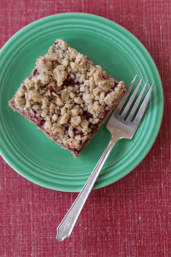 A raspberry bar with a rich, buttery crust and a crispy crumble topping, placed on a green plate with a fork on the left side.
