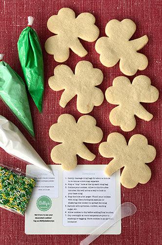 Six clover-shaped sugar cookies and a DIY cookie decorating kit on a red background. The kit includes green and white icing bags with tips, and a bag of green and white sprinkles, alongside a white instruction card from 'Billy's' bakery.