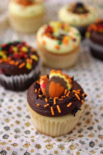 Thanksgiving cupcakes decorated with chocolate frosting, orange sprinkles, and a candy turkey on a white polka-dot tablecloth.