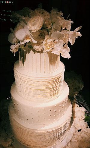 Elegant white four-tiered wedding cake decorated with ruffled icing, pearl accents, and topped with white roses and orchids.