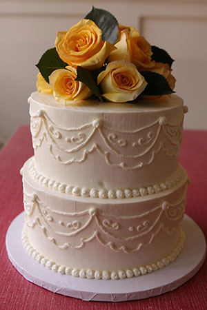 Two-tier white wedding cake decorated with white piping and topped with yellow roses.