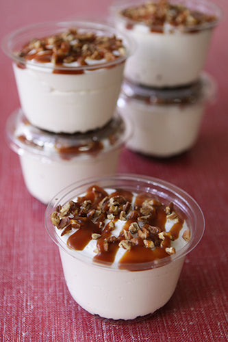 Five individual servings of turtle cheesecake in clear plastic cups, topped with a drizzle of caramel sauce and chopped pecans.