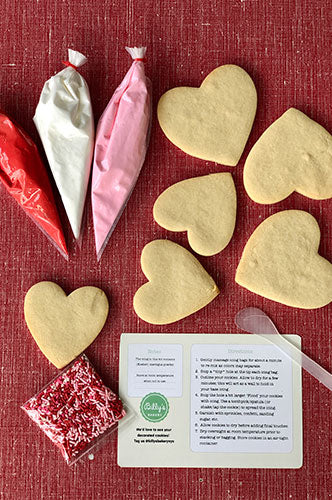 The image shows a Valentine's cookie decorating kit which includes six sugar heart cookies, three royal icing pouches, one garnish pouch, and a mini spatula. The background is a red textured surface.