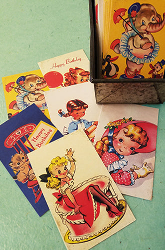 A collection of vintage birthday cards featuring illustrations of cartoon children and animals, including elephants and kittens, with text "Happy Birthday" visible on some cards. The cards are arranged on a light-colored surface next to a metal cardholder containing more cards.